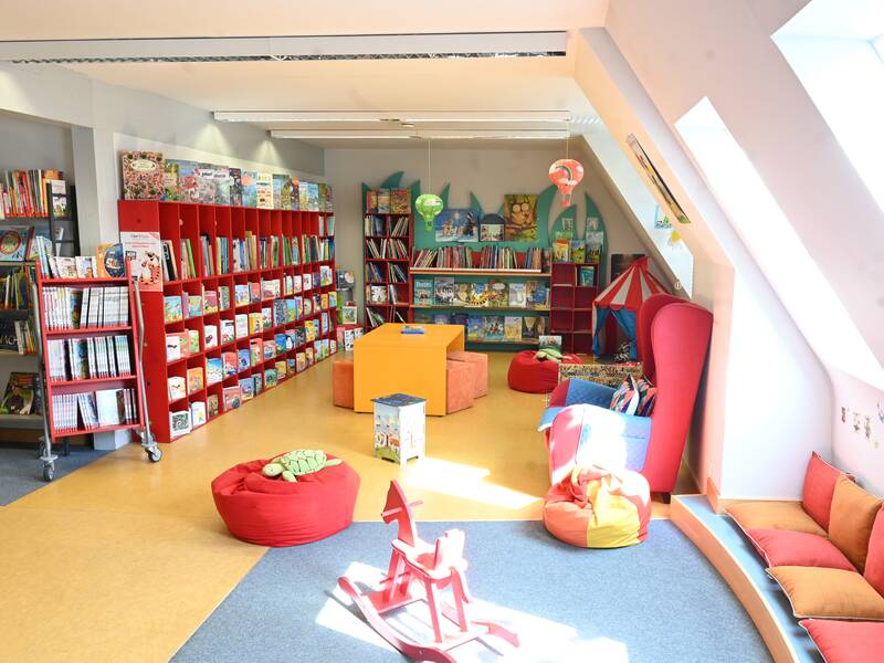 Ein bunter, einladender Raum einer Kinderbibliothek mit roten Bücherregalen, verschiedenen Kinderbüchern, Sitzsäcken und einem kleinen Zelt.