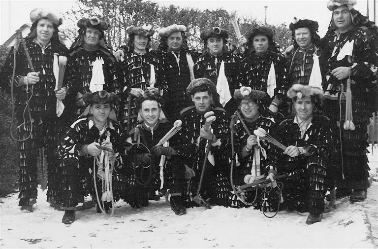 Urzeln / Eine Gruppe von Menschen trägt traditionelle, auffällige Kostüme mit Fellmützen und hält verschiedene Gegenstände, während sie draußen im Schnee posieren.