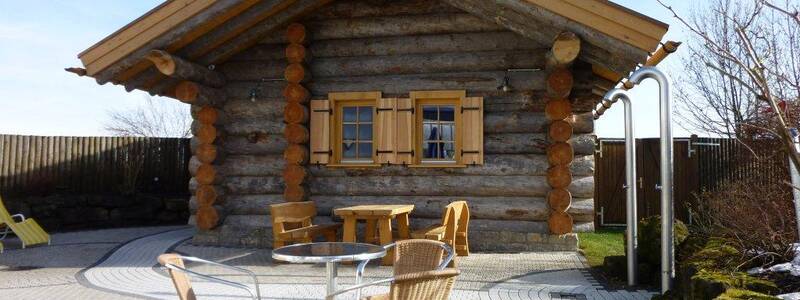 Das Bild zeigt ein Holzhaus von vorne, im Vordergrund stehen Stühle und ein Tisch.