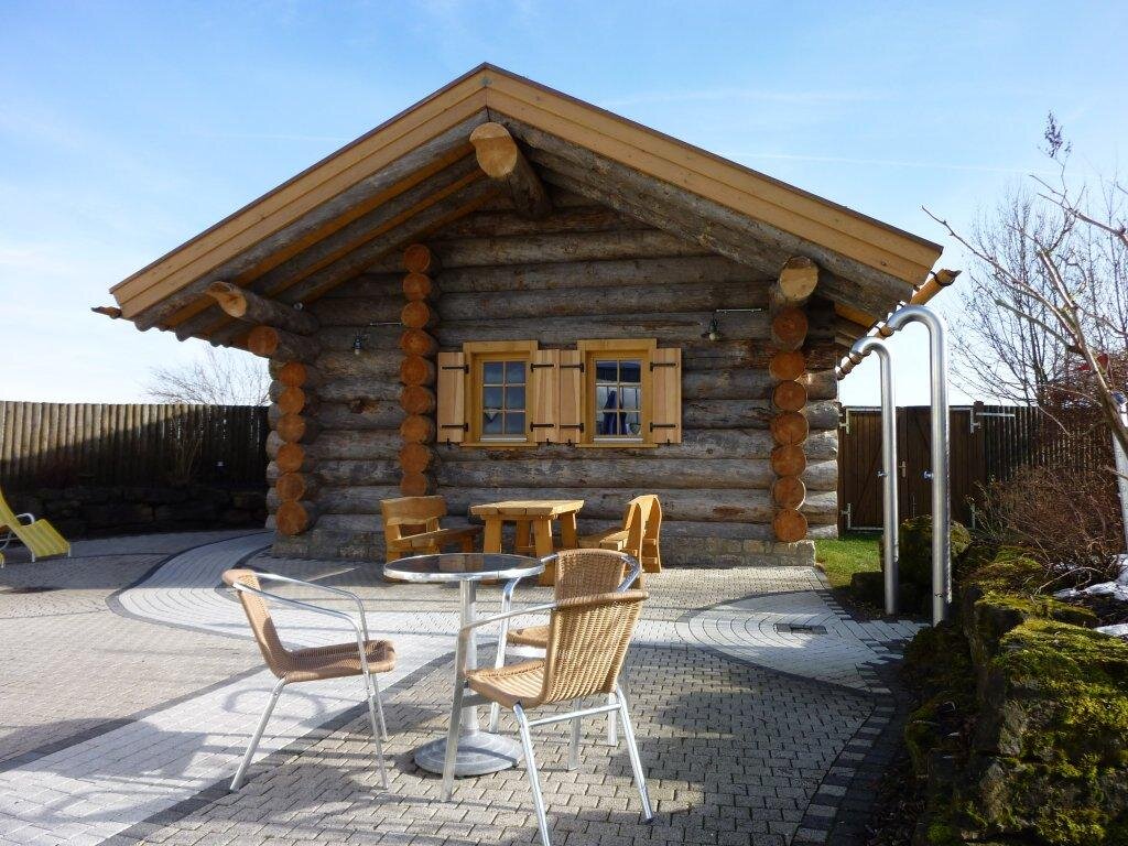 Das Bild zeigt ein Holzhaus von vorne, im Vordergrund stehen Stühle und ein Tisch.