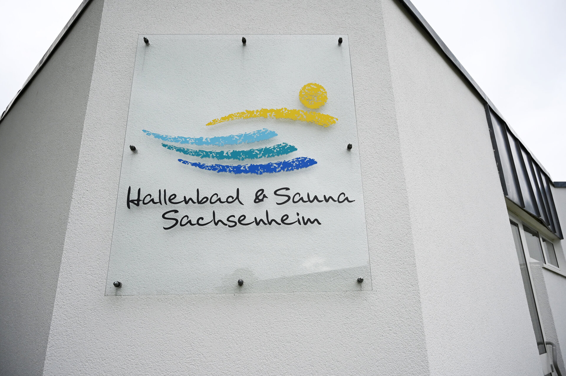 Ein Schild mit der Aufschrift "Hallenbad & Sauna Sachsenheim" ist an einer weißen Gebäudewand angebracht, verziert mit stilisierten blauen und gelben Wellen und einer Sonne.