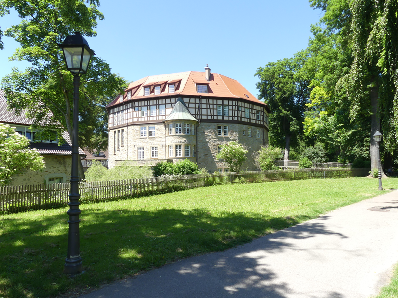 Das Wasserschloss mit rotem Dach steht in einem grünen, parkähnlichen Umfeld mit Laternen und Bäumen.