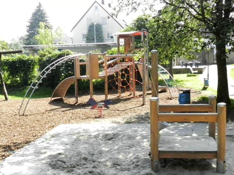 Der Spielplatz zeigt eine Kletterstruktur aus Holz und Metall mit Netzen und Rutschen sowie eine federnde Wippe im Vordergrund, umgeben von Bäumen und Sand.