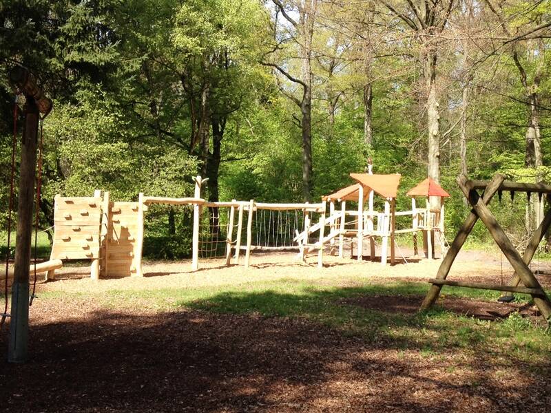 Ein hölzerner Abenteuerspielplatz mit Kletterwand und Spinnennetzbrücke steht in einem waldigen Bereich im Sonnenlicht.