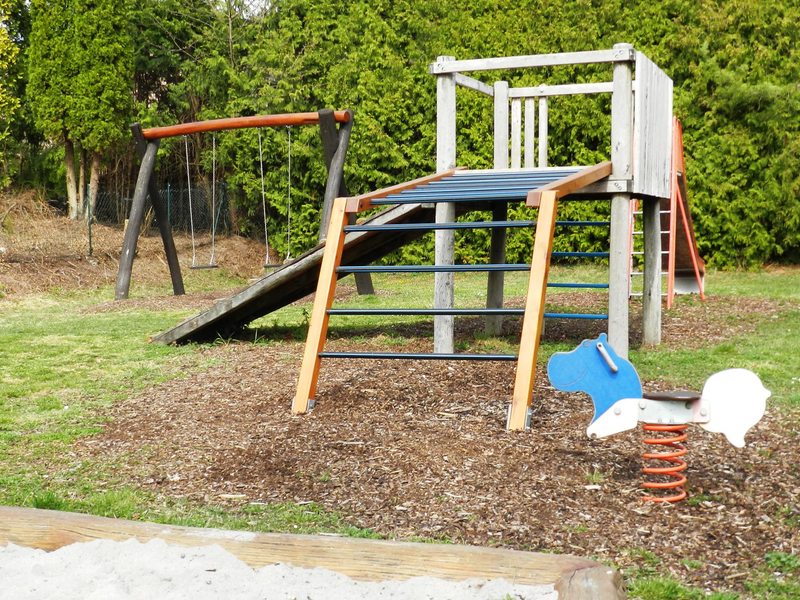 Ein Spielplatz mit Klettergerüst, Rutsche, Schaukel und Wipptier befindet sich auf einer Wiese vor einer Hecke.