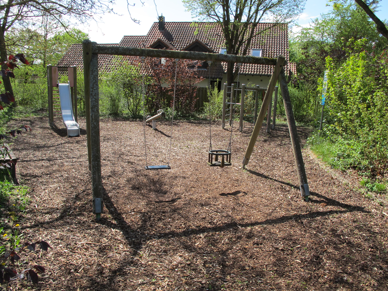 Ein kleiner Spielplatz mit einer Schaukel, einer Rutsche und einem Wipptier, umgeben von Bäumen und Büschen, liegt vor einem Haus mit Ziegeldach.