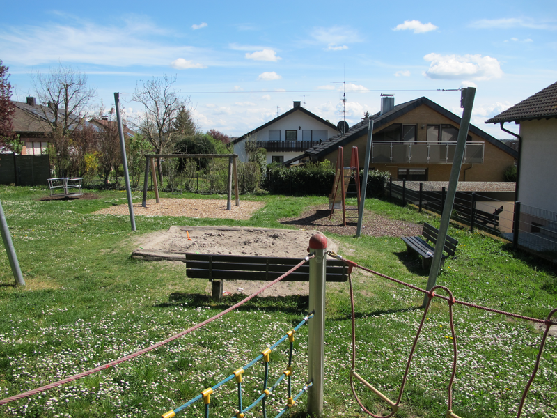Ein Spielplatz mit Klettergerüst, Schaukeln, einem Sandkasten und Sitzbänken, umgeben von grüner Wiese und Wohnhäusern im Hintergrund.