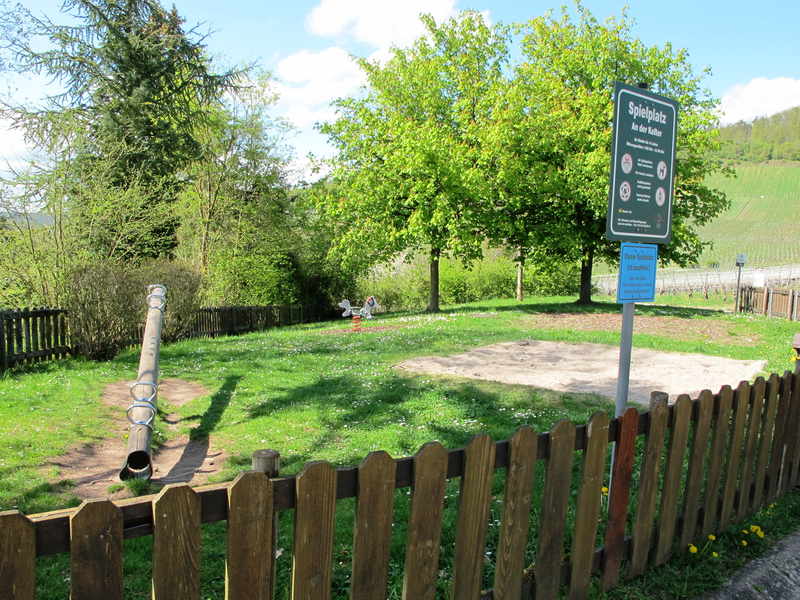 Ein umzäunter Spielplatz im Grünen mit einer Wippe, einem Sandkasten und einem Schild mit Informationen.