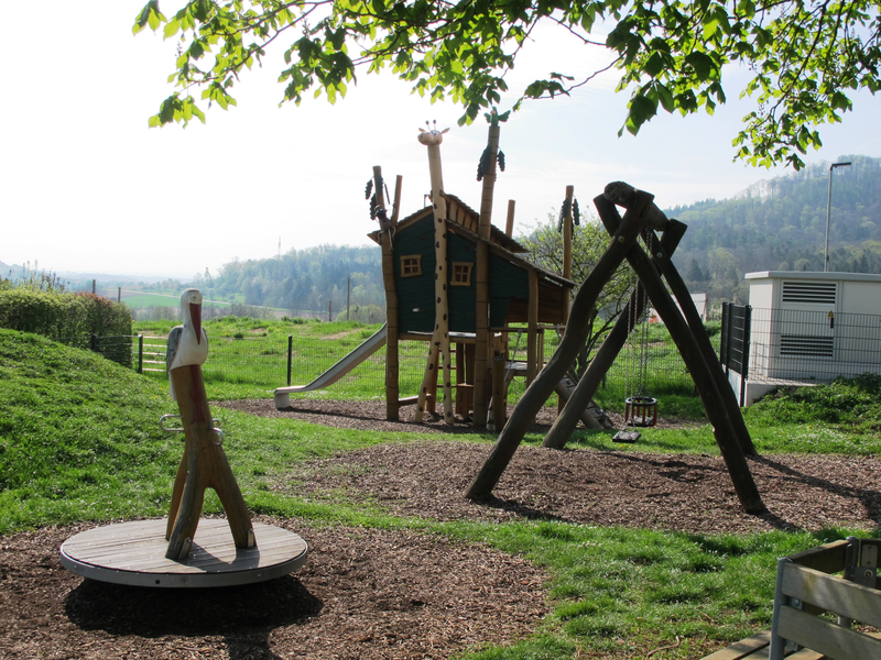 Ein Spielplatz mit einer Holzwippe in Form eines Vogels, einer Holzschaukel und einem kleinen Spielhaus, umgeben von grünen Hügeln und Bäumen.