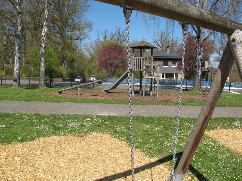 Ein Spielplatz mit einer Schaukel, Rutsche und Klettergerüst, umgeben von Bäumen und einem Haus im Hintergrund.