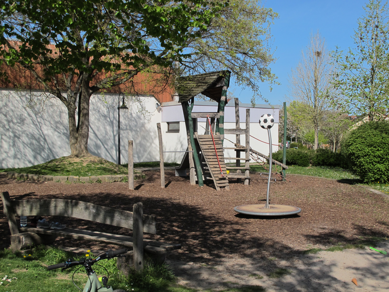 Ein Spielplatz mit Klettergerüst und Karussell, umgeben von Bäumen und Rasenfläche bei sonnigem Wetter.