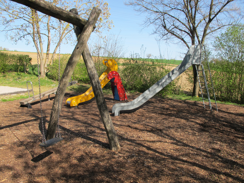 Ein Spielplatz mit einer Schaukel, einer gelben Rutsche in Form eines Tieres und einer großen Metallrutsche, umgeben von Bäumen.