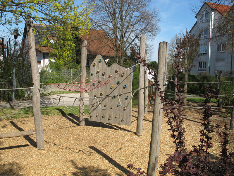 Ein Spielplatz mit einer Kletterwand, Seilen und Holzpfosten, umgeben von Bäumen und Häusern im Hintergrund.