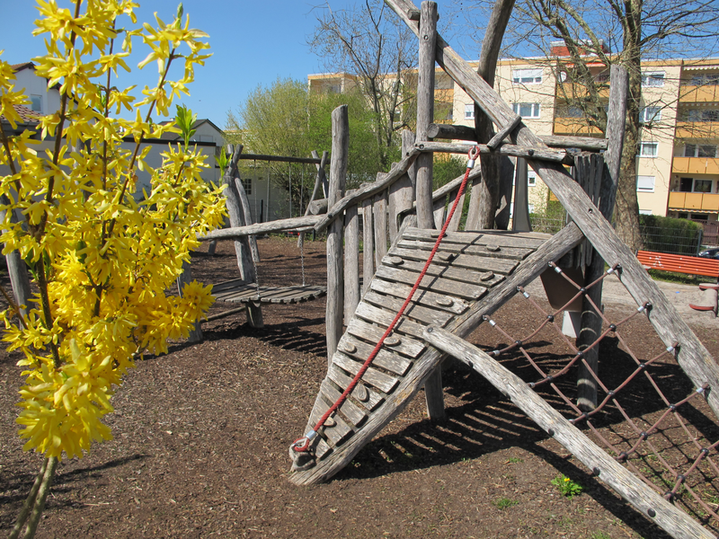 Ein Spielplatz aus Holz mit Klettergerüsten und einem gelb blühenden Strauch im Vordergrund, umgeben von Wohngebäuden.