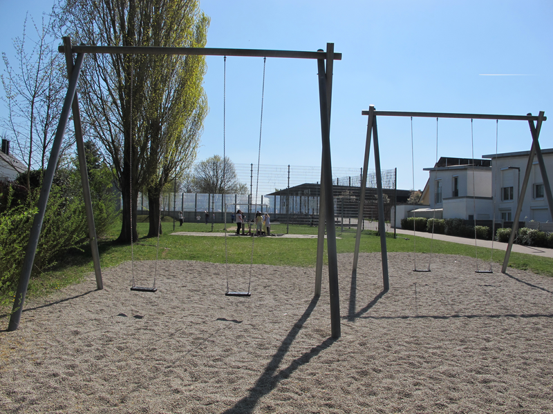 Eine leere Schaukel aus Holz steht auf einem Spielplatz mit Rasen und Bäumen im Hintergrund.