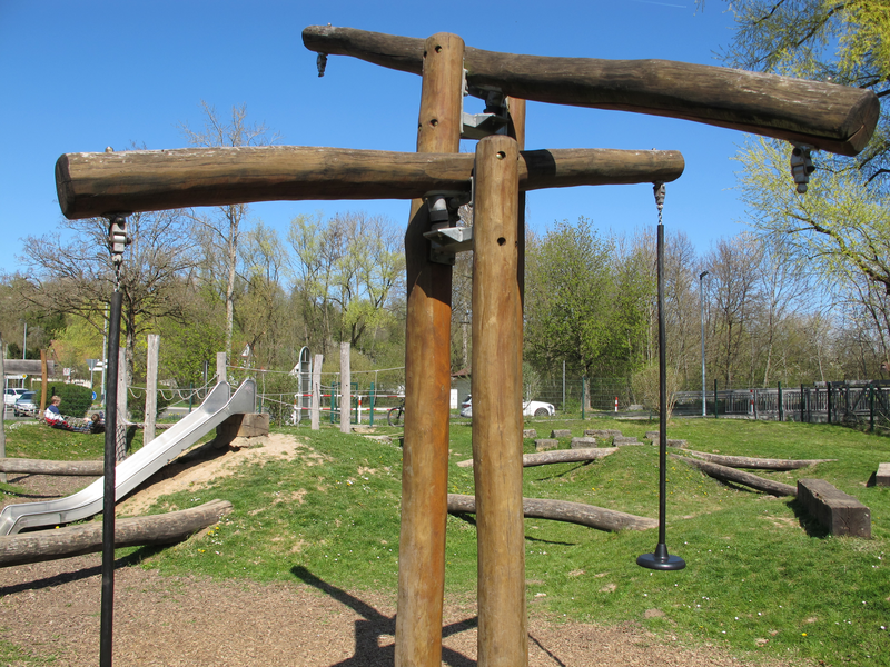 Ein Spielplatz mit einer Rutsche, Holzkonstruktionen und grüner Wiese im Hintergrund.
