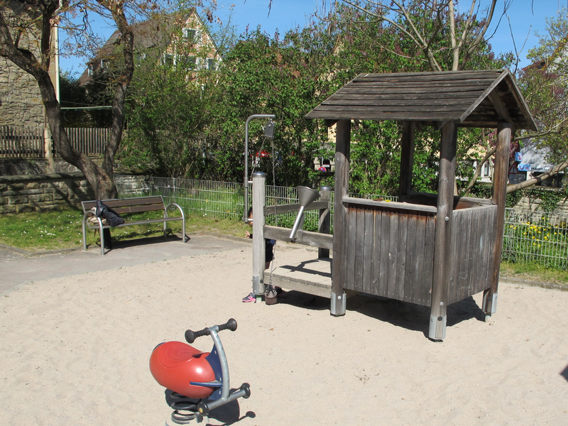 Ein Spielplatz mit einem kleinen Holzhaus auf einer Sandfläche, einer roten Wippe und einer Holzbank im Hintergrund.