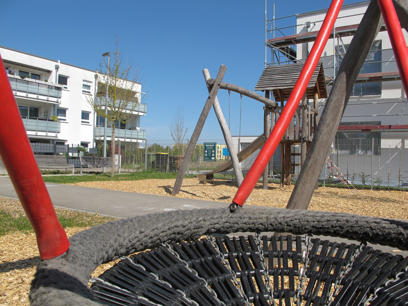 Ein Spielplatz mit einem modernen Klettergerüst und einer Schaukel befindet sich in einer Wohngegend mit mehrstöckigen Gebäuden im Hintergrund.
