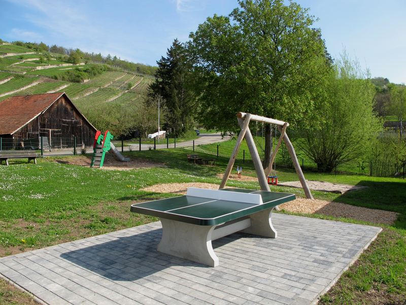Ein Spielplatz im Grünen mit einer Tischtennisplatte im Vordergrund, einer Schaukel und einer Rutsche neben einem Bauernhof und terrassierten Hügeln im Hintergrund.