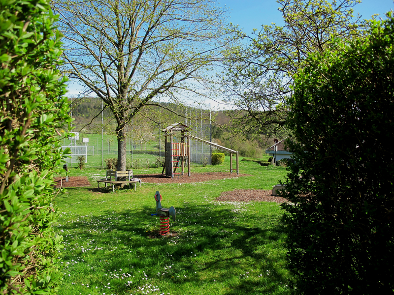 Ein grüner Spielplatz mit Bäumen, Klettergerüst, Wippe und Tischtennisplatte ist von Hecken umgeben.