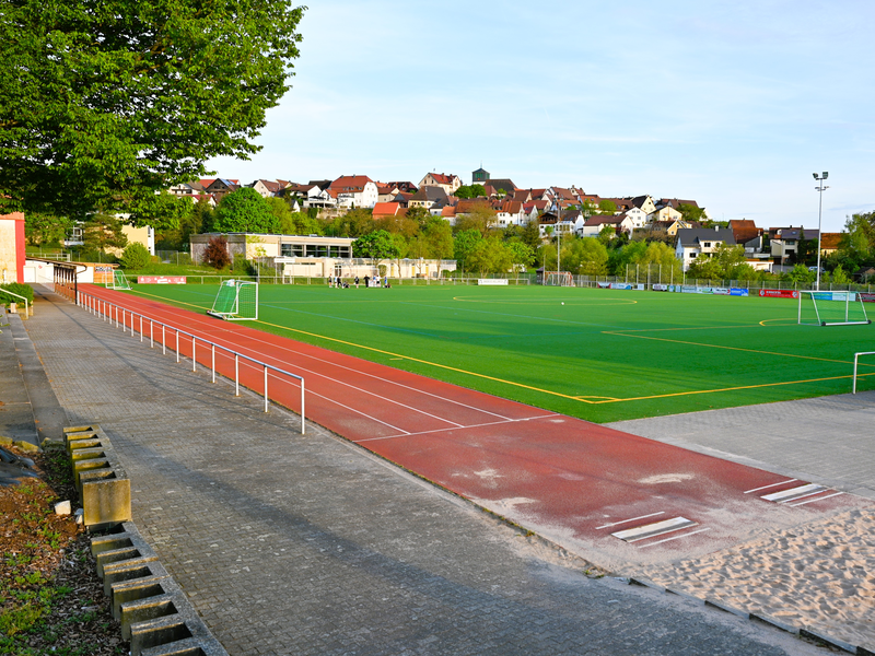 Ein grüner Fußballplatz mit umliegender Laufbahn liegt vor einer Reihe von Wohnhäusern auf einem Hügel, umgeben von Bäumen.