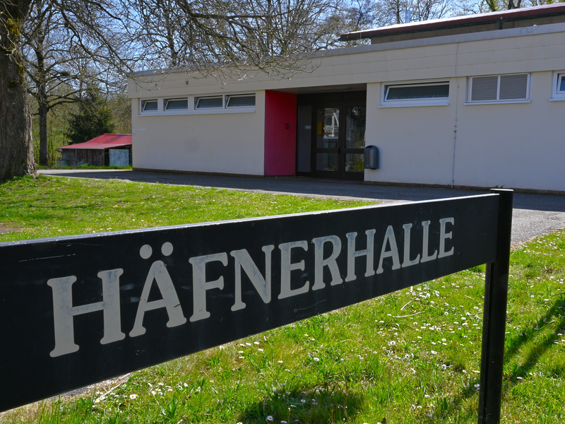 Ein flaches Gebäude mit weißen Wänden und einem roten Eingang ist hinter einem Schild mit der Aufschrift "Häfnerhalle" zu sehen.