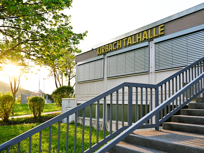 Das Bild zeigt ein Gebäude mit der Aufschrift "KIRBACHTALHALLE" an der Fassade, umgeben von grünen Bäumen und Rasenflächen im Sonnenuntergang.