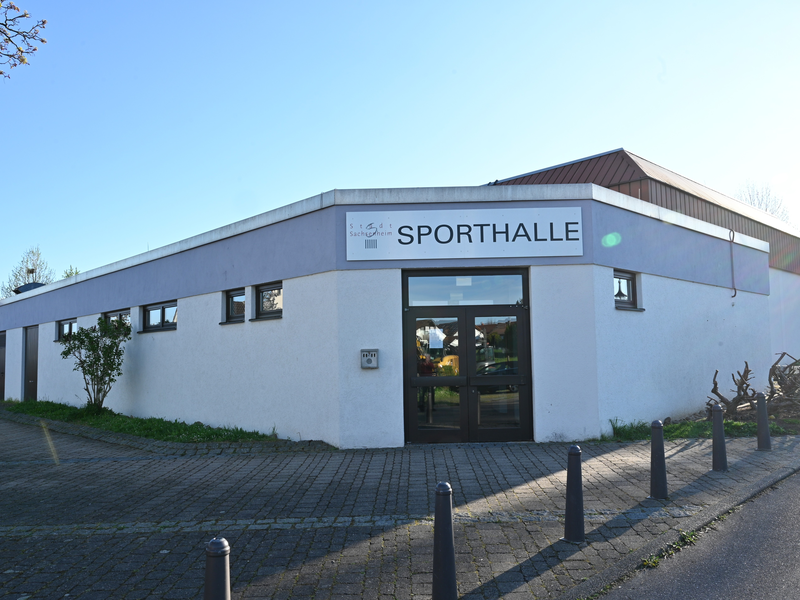 Ein flaches Gebäude mit der Aufschrift "SPORTHALLE" über dem Eingang steht bei sonnigem Wetter an einem gepflasterten Weg.