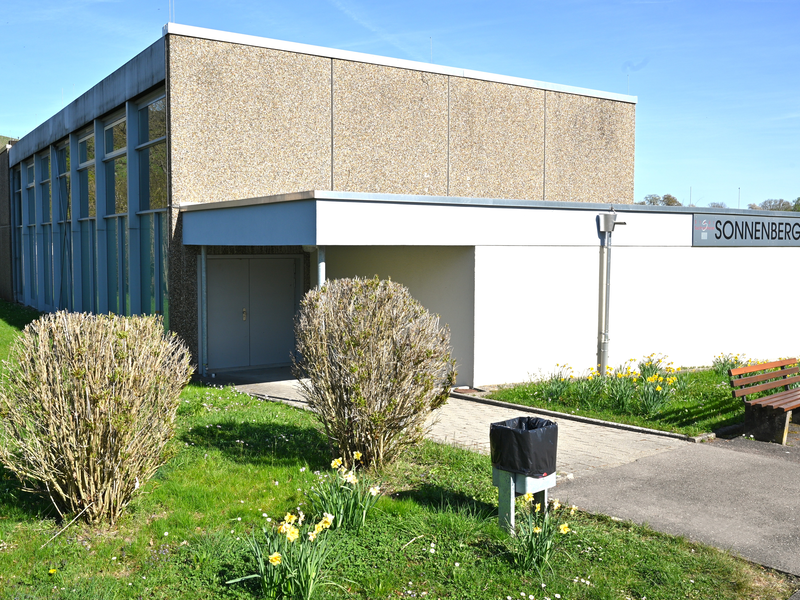 Das Bild zeigt ein modernes Gebäude mit großer Fensterfront, Beschilderung "Sonnenberghalle" und einem Weg, umgeben von gepflegtem Rasen und blühenden Sträuchern.
