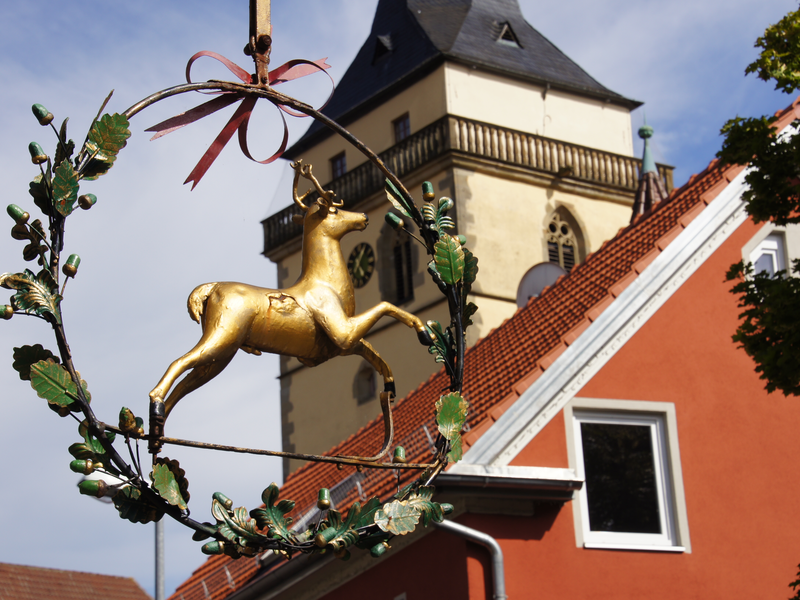 Ein goldenes Hirschornament hängt vor einem rot gedeckten Gebäude und einem Kirchturm.