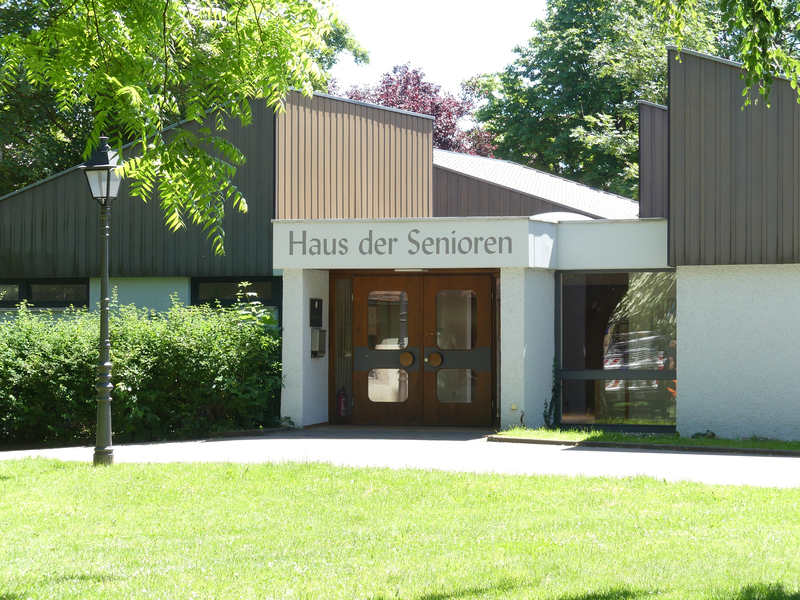 Das Bild zeigt den Eingang eines Gebäudes mit der Aufschrift "Haus der Senioren", umgeben von Bäumen und Grünflächen.
