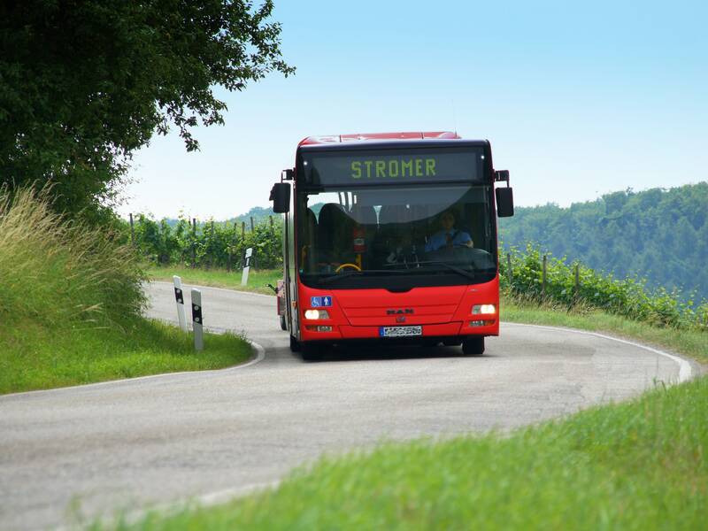 Ein roter Bus mit der Aufschrift "STROMER" fährt auf einer kurvigen Landstraße durch eine grüne Landschaft.