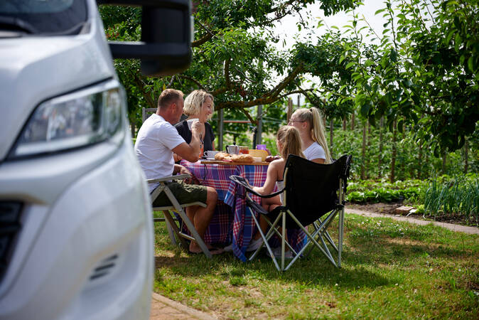 Eine Familie sitzt an einem Tisch im Freien, genießt eine Mahlzeit unter Bäumen, während ein weißes Auto im Vordergrund am Rand sichtbar ist.