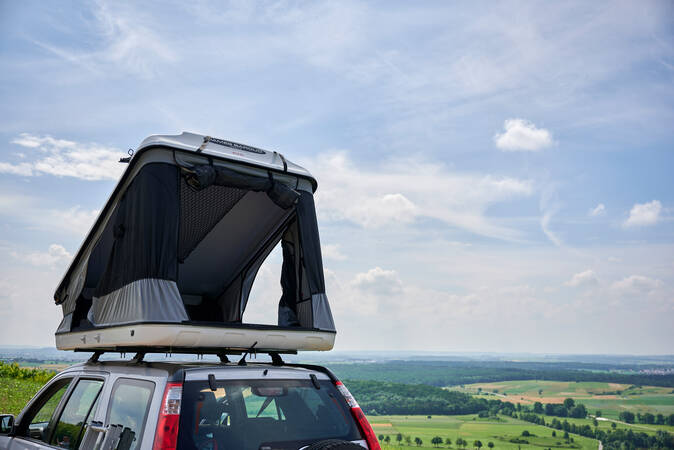Ein Auto mit einem geöffneten Dachzelt steht vor einer weiten, grünen Landschaft unter einem teilweise bewölkten Himmel.