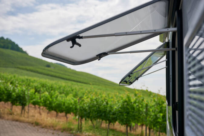 Die Nahaufnahme zeigt ein Wohnmobilfenster, das geöffnet ist, mit einer Landschaft aus Weinbergen im Hintergrund.
