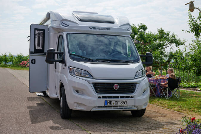 Ein weißes Wohnmobil steht auf einem Parkplatz mit geöffneter Tür, und im Hintergrund sitzt eine Familie an einem Tisch im Freien.