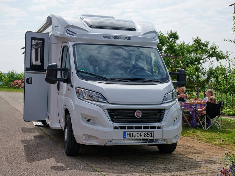 Ein weißes Wohnmobil steht auf einem Parkplatz mit geöffneter Tür, und im Hintergrund sitzt eine Familie an einem Tisch im Freien.