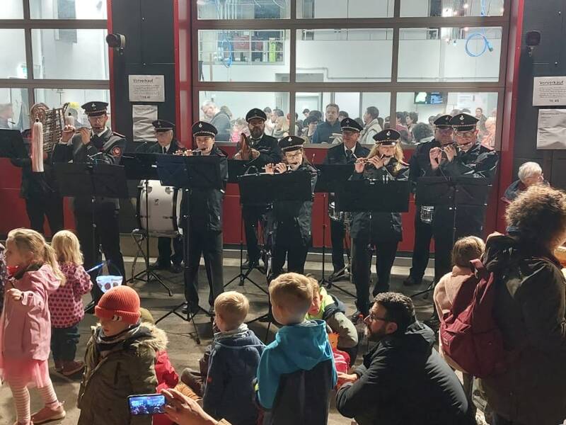 Eine Blaskapelle in Uniformen spielt vor einem Publikum, das aus Erwachsenen und Kindern besteht, in einer belebten Innenraumszene.