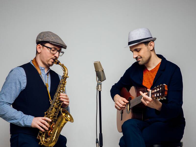 Zwei Männer musizieren zusammen, einer spielt Saxophon und der andere Gitarre, vor einem Mikrofon.