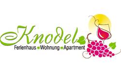 Das Bild zeigt das Logo eines Ferienhausanbieters mit dem Schriftzug "Knodel Ferienhaus Wohnung Apartment" in grün und schwarz, ergänzt durch eine Weintraube und ein Weinglas vor einer gelben Sonne.