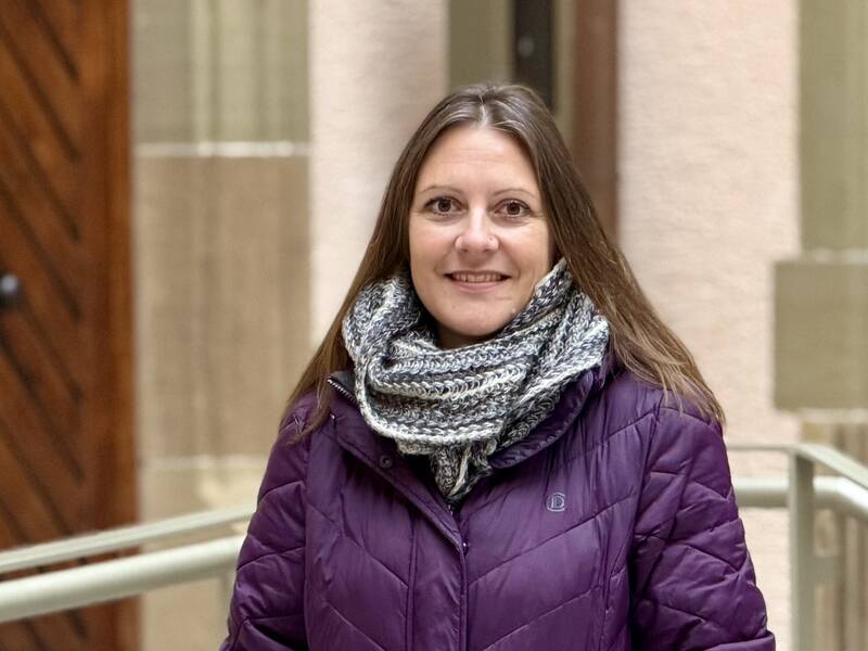 Eine Frau mit langen Haaren trägt eine lilafarbene Jacke und einen grauen Schal, während sie in einer Umgebung mit Holztür und Geländer steht.