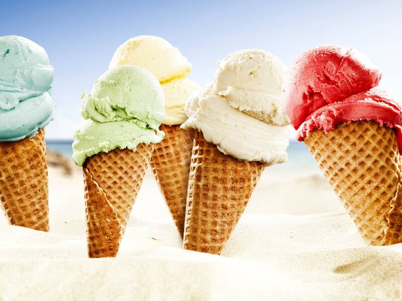 Fünf Eiswaffeln mit verschiedenfarbigem Eis stehen im Sand vor einem verschwommenen Hintergrund, der an einen Strand erinnert.