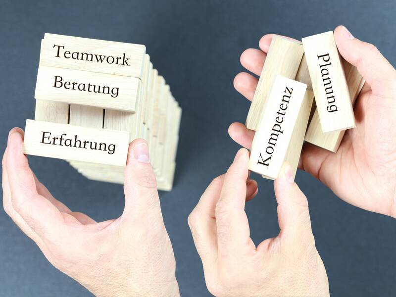 Zwei Hände halten Holzblöcke mit den Wörtern "Teamwork", "Beratung", "Erfahrung", "Kompetenz" und "Planung" vor einem dunkelgrauen Hintergrund.