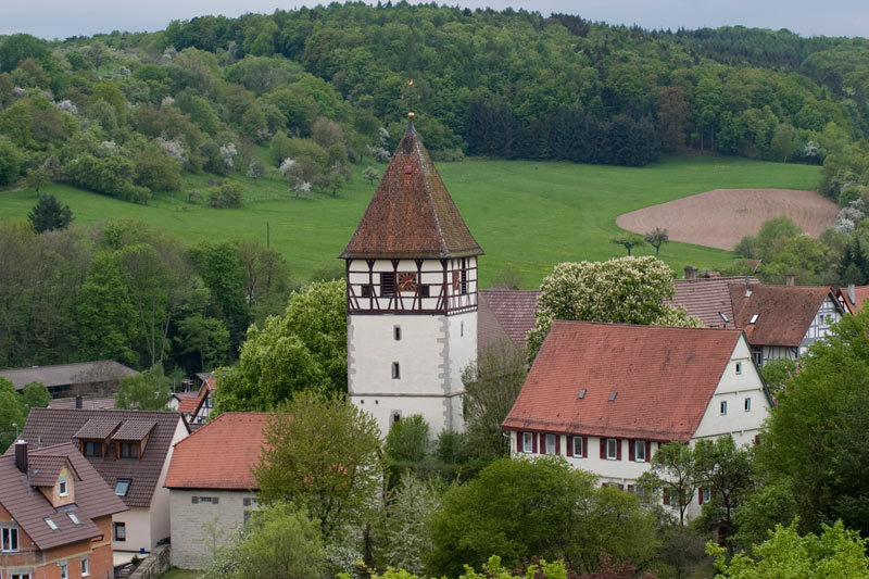 Ein malerisches Dorf mit einem historischen Turm, umgeben von üppigen grünen Hügeln und Wäldern.